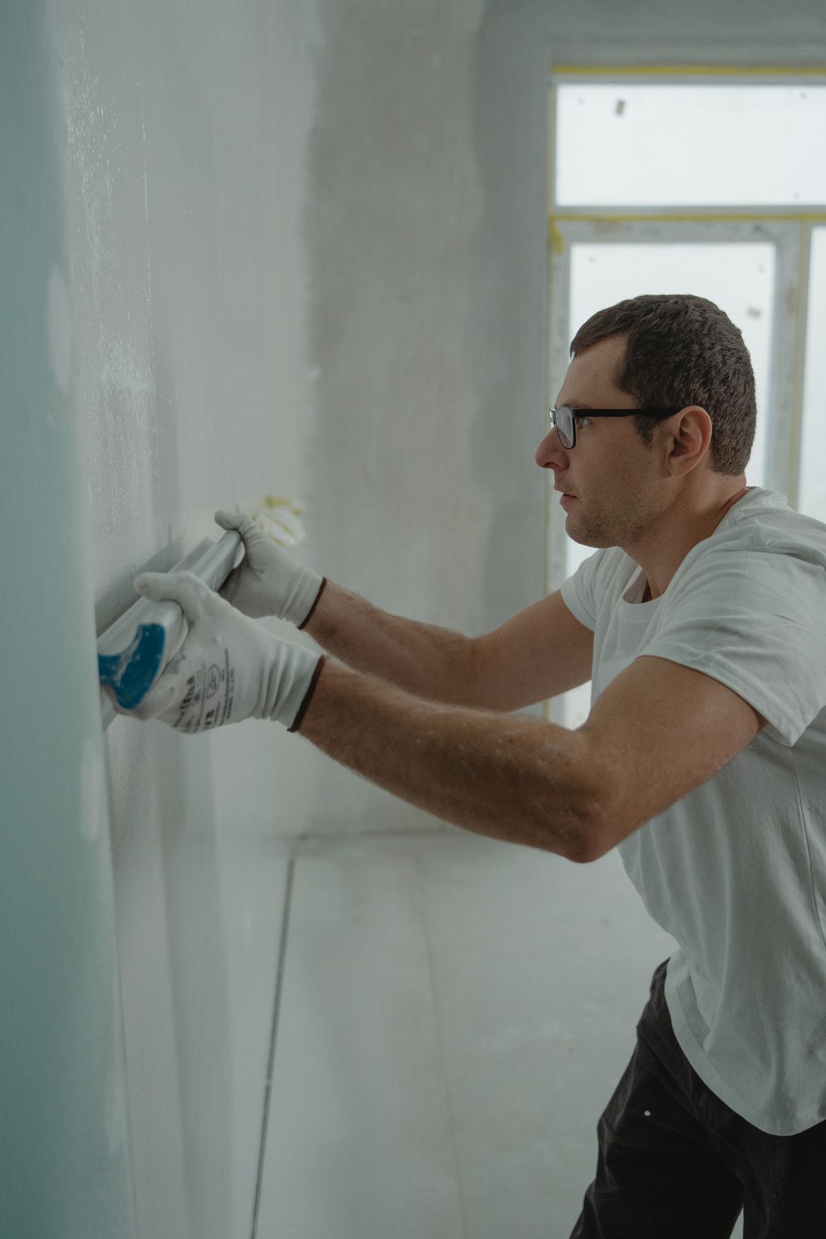 Gloved worker smoothing fresh joint compound onto a drywall surface with a taping knife